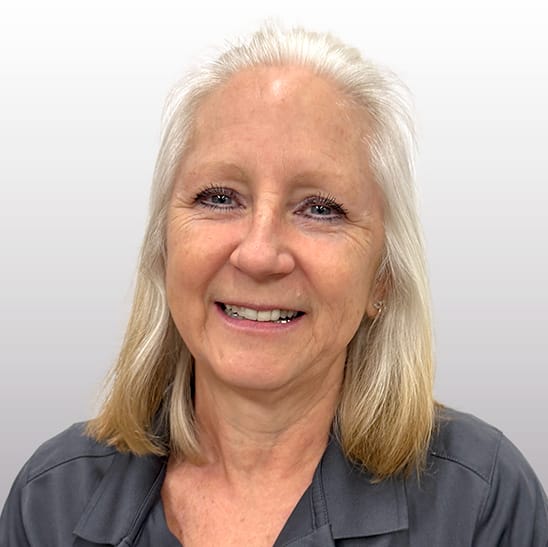 Older woman with straight, shoulder-length gray hair, wearing a gray collared shirt, smiling in front of a light gray background. Older woman with straight, shoulder-length gray hair, wearing a gray collared shirt, smiling in front of a light gray background.