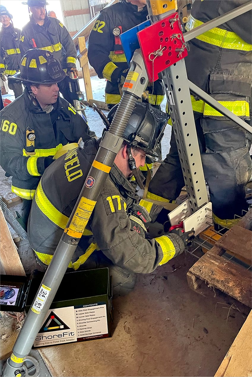 Firefighters in protective gear adjust a stabilization device on a structure inside a building, with one firefighter kneeling and others observing.